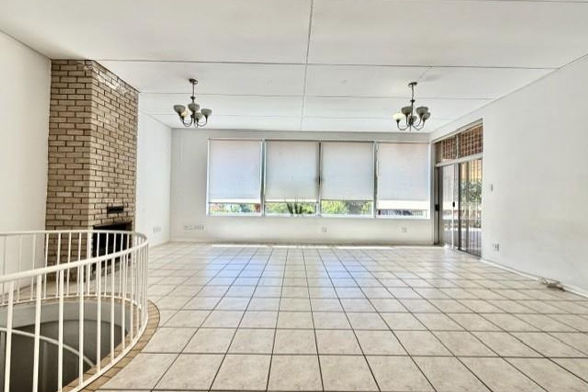 Spacious open-plan living area featuring a brick fireplace, tiled flooring, and large windows with roller blinds, leading to an outdoor patio area.