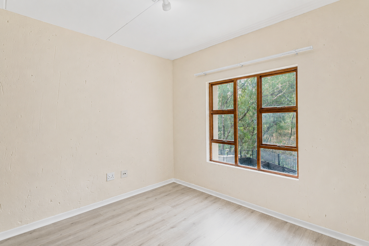 An empty bedroom featuring light-toned laminate flooring, textured walls, and a wooden-framed window overlooking greenery.