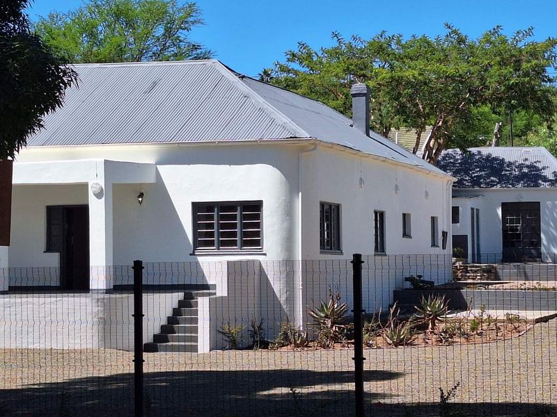A well-maintained guest house exterior featuring white rendered walls, a corrugated metal roof, and a secure fenced perimeter.