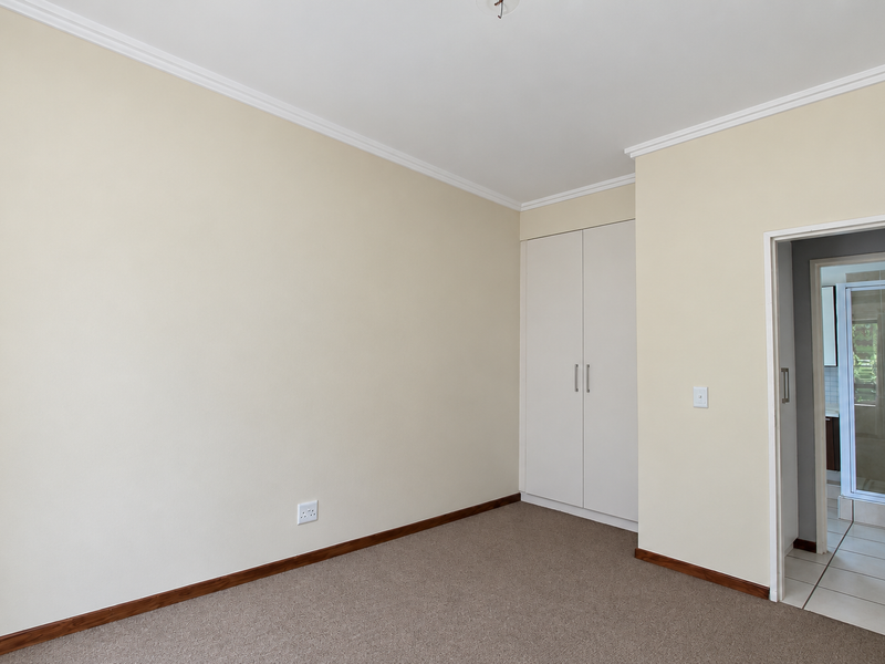 A clean, empty bedroom featuring neutral walls, carpeted flooring, and built-in cupboards, with an adjacent en-suite bathroom visible through the doorway.