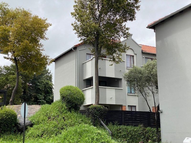 Exterior view of a multi-story apartment building featuring balconies, plastered walls, and a tiled roof, set within a landscaped residential complex.