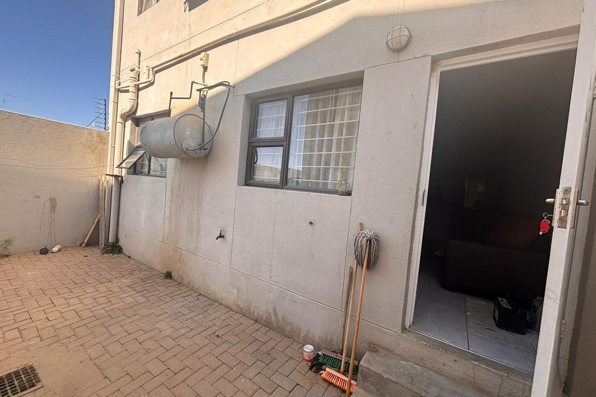 A rear courtyard area featuring paved flooring, an external geyser, and security bars on the windows, providing access to the apartment interior.