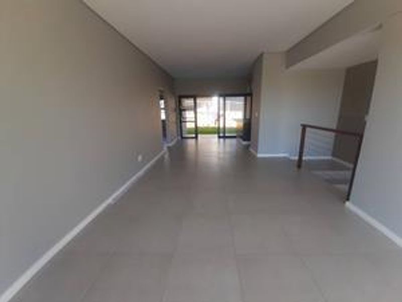 A spacious, empty open-plan living area featuring neutral tiled flooring, modern wall finishes, and a staircase railing leading to an outdoor patio.