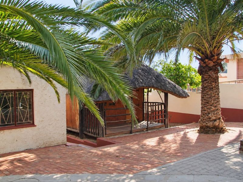 A rear garden area featuring a thatched lapa structure, brick paved courtyard, and mature palm tree, enclosed by a boundary wall.