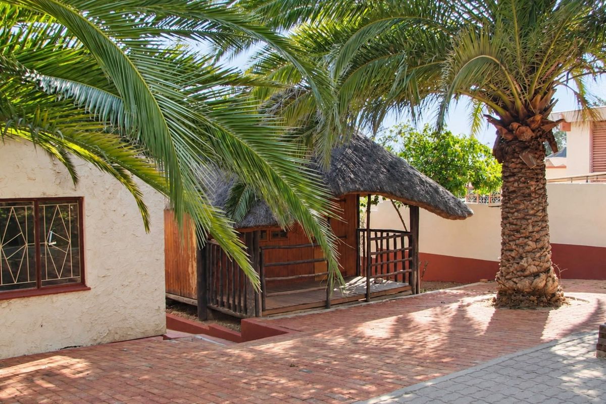 A rear garden area featuring a thatched lapa structure, brick paved courtyard, and mature palm tree, enclosed by a boundary wall.