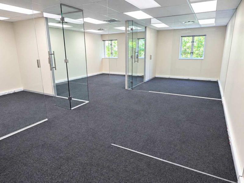 A bright, modern open-plan office space featuring glass partitions, suspended ceilings with recessed lighting, and clean carpet flooring.