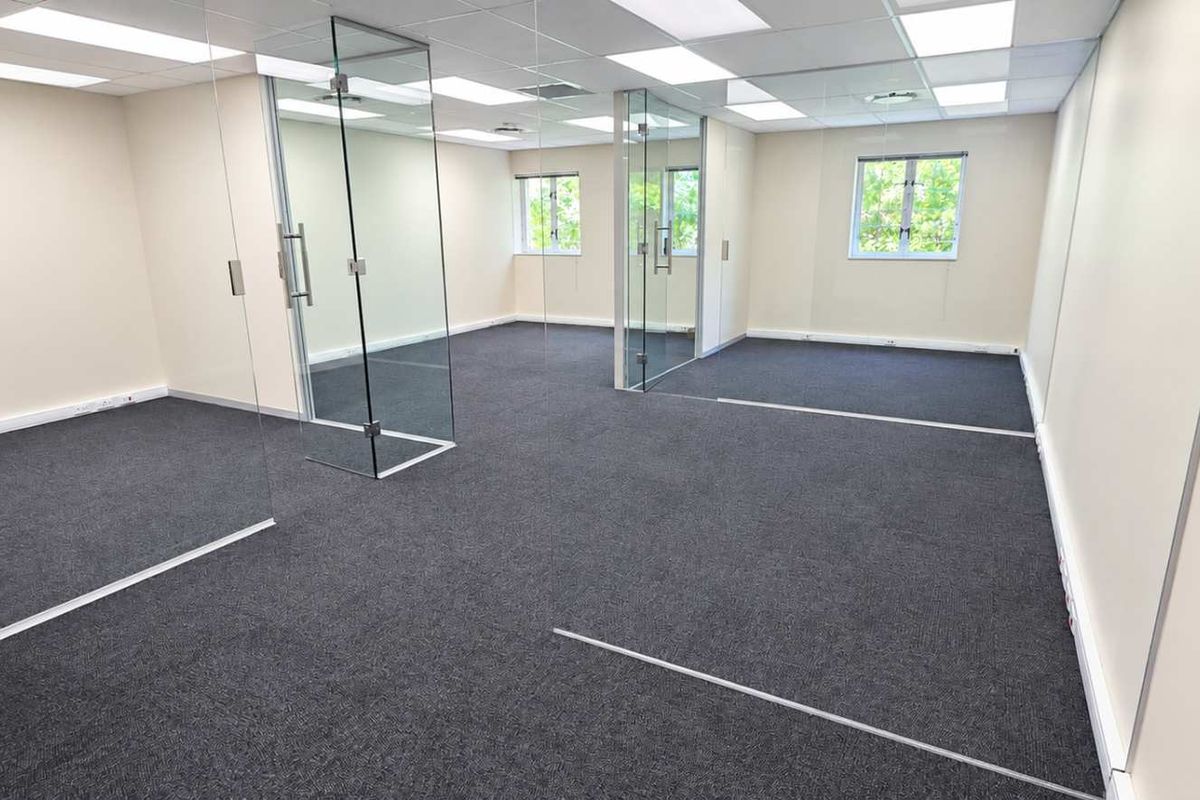 A bright, modern open-plan office space featuring glass partitions, suspended ceilings with recessed lighting, and clean carpet flooring.
