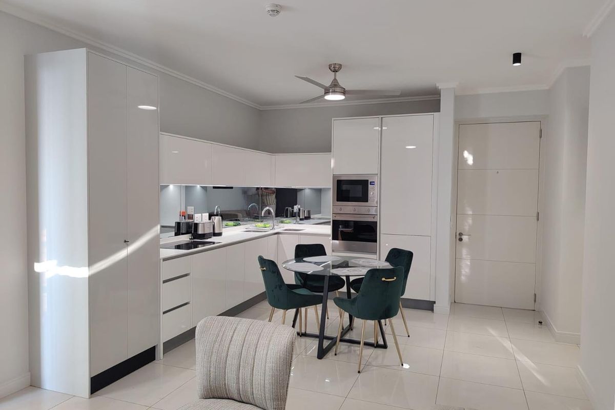 Modern open-plan kitchen featuring high-gloss white cabinetry, integrated appliances, mirror splashback, and sleek tiled flooring in a bright, contemporary living space.
