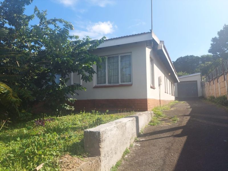 Front exterior view of the house featuring a driveway, concrete retaining wall, and a detached garage, showing signs of wear and overgrown landscaping.