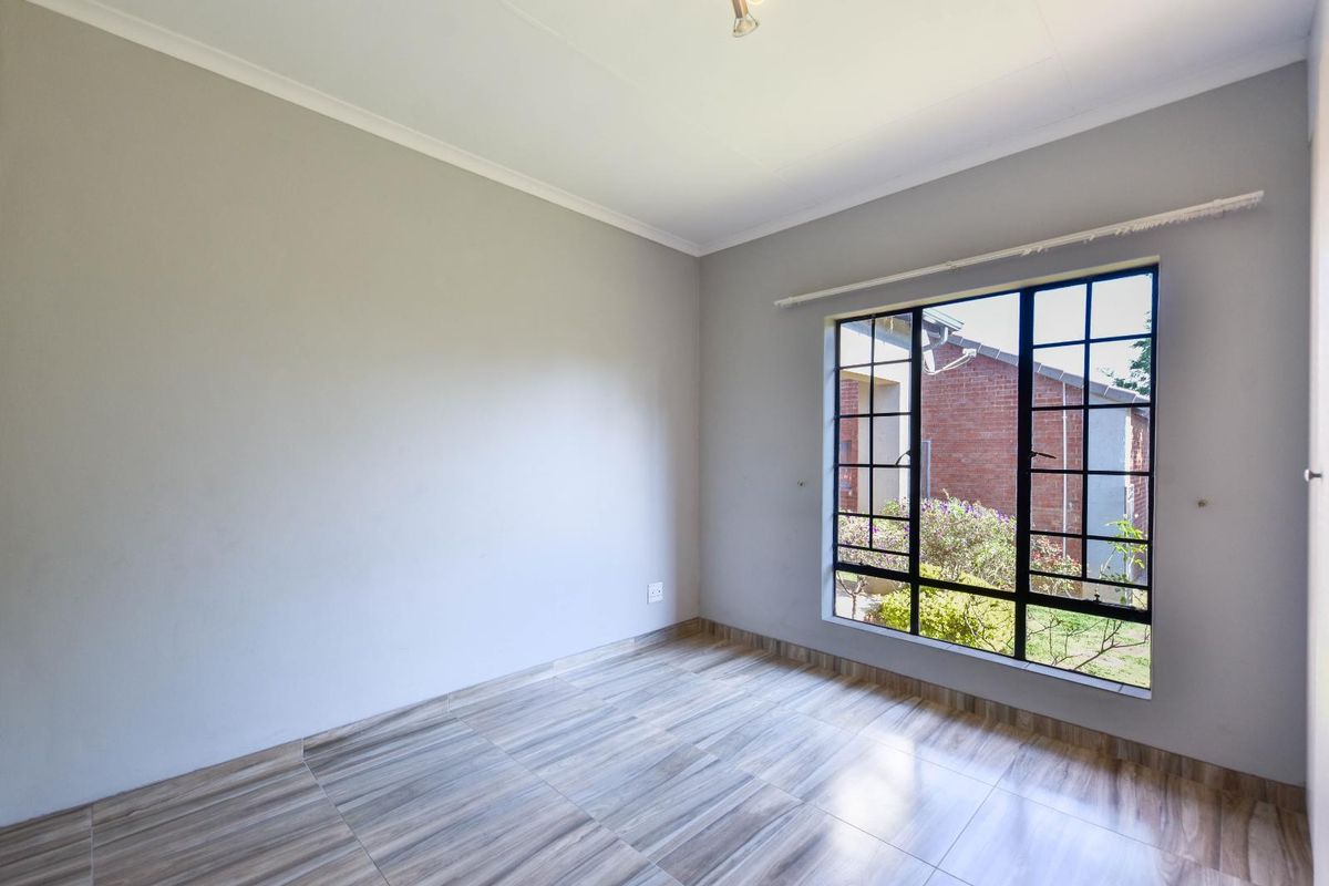 A bright, empty bedroom featuring modern wood-look tiled flooring, neutral wall paint, and a window fitted with security burglar bars.