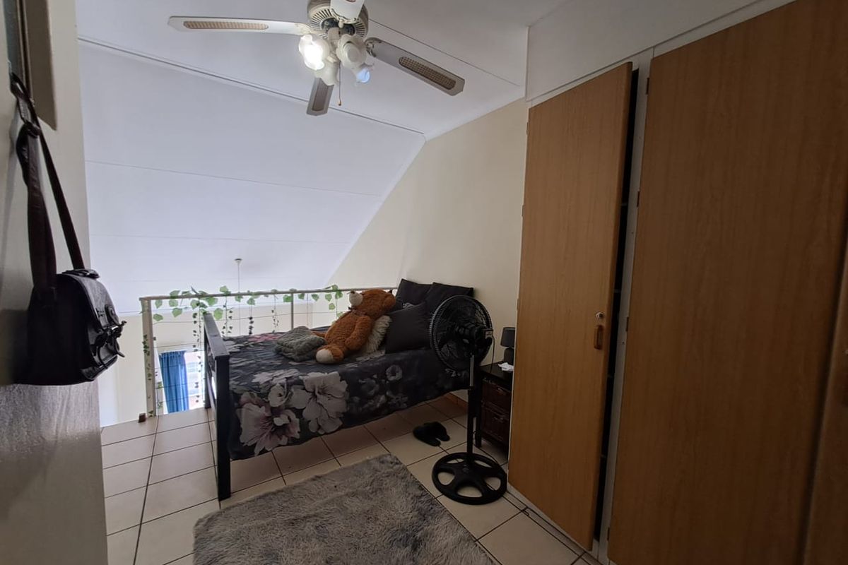 A loft-style bedroom featuring tiled flooring, built-in wooden cupboards, and a ceiling fan, overlooking the lower level via a mezzanine railing.
