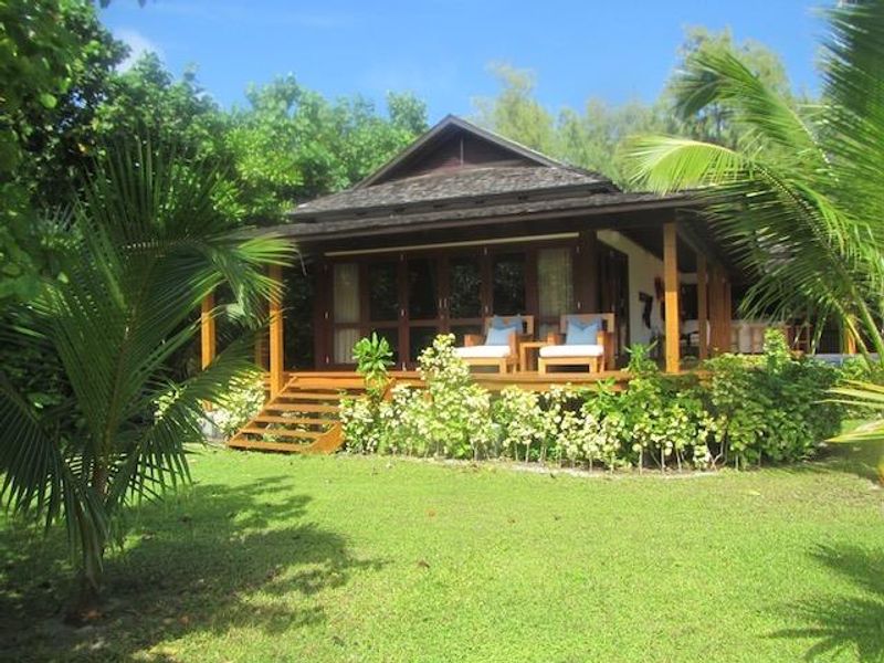 A charming villa exterior featuring a wooden deck, covered patio area, and lush tropical garden surroundings.