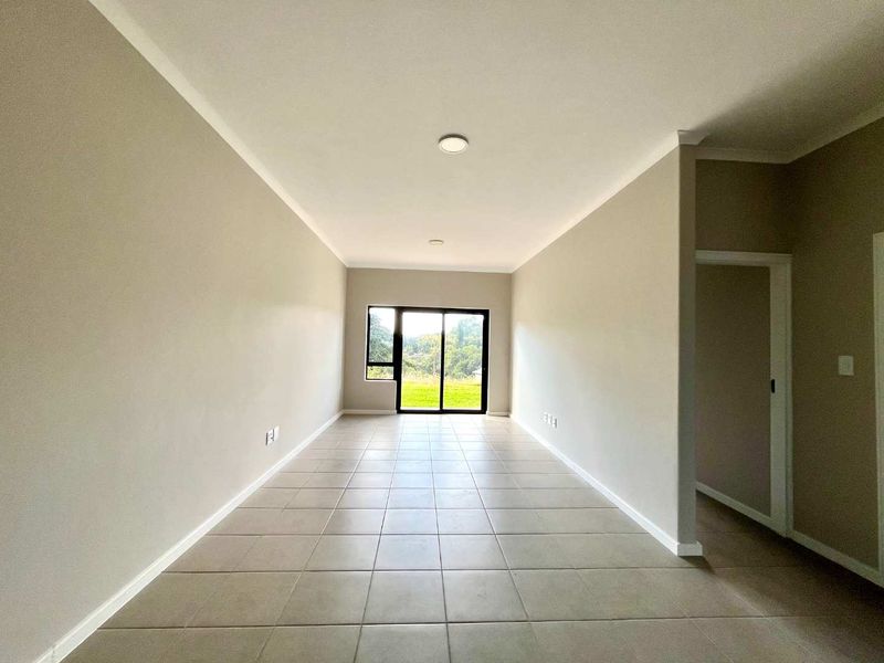 A bright, empty living area featuring neutral walls, tiled flooring, and a sliding glass door providing natural light and garden views.