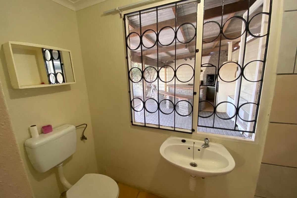 A functional bathroom featuring a wall-mounted basin, toilet, and a window with security bars overlooking an outdoor patio area.