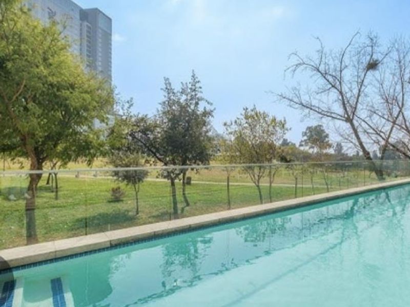 A clear swimming pool featuring a modern glass balustrade, overlooking a landscaped green area and urban surroundings.