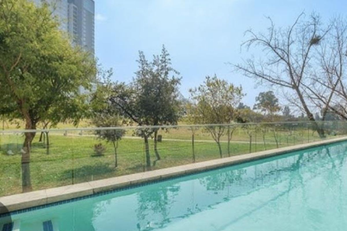 A clear swimming pool featuring a modern glass balustrade, overlooking a landscaped green area and urban surroundings.