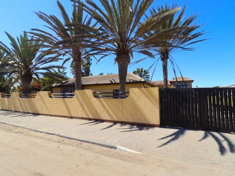 Street view of the property featuring a yellow boundary wall, dark timber gate, and mature palm trees along the roadside.