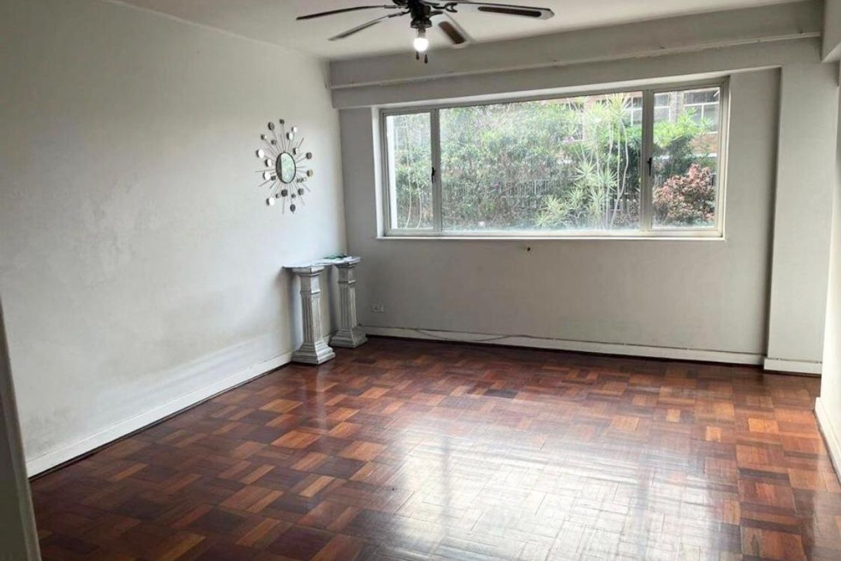 A spacious, empty living room featuring classic parquet flooring, a ceiling fan, and a large window providing natural light.