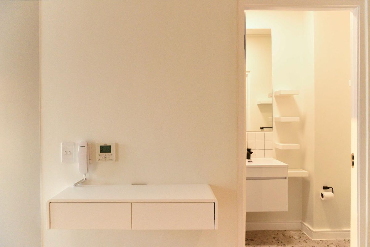 A clean, modern bathroom featuring a wall-mounted vanity, floating shelving, and neutral tiled flooring, adjacent to a hallway with an intercom and thermostat.
