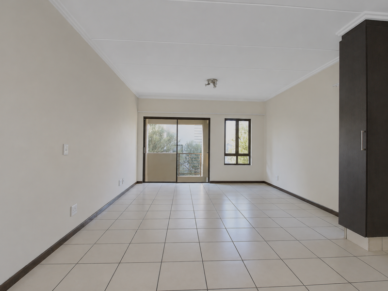 A bright, open-plan living area featuring neutral tiled flooring, built-in dark cabinetry, and access to a balcony through a sliding glass door.