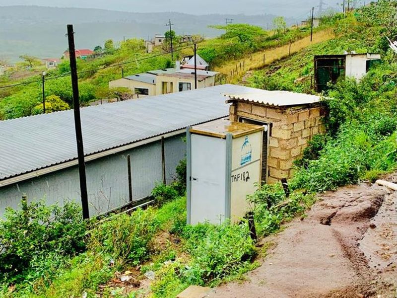 An exterior view of a sloped property area featuring a corrugated metal roof structure, a utility box, and an unpaved, eroded dirt path.