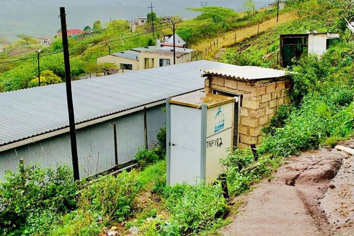 An exterior view of a sloped property area featuring a corrugated metal roof structure, a utility box, and an unpaved, eroded dirt path.