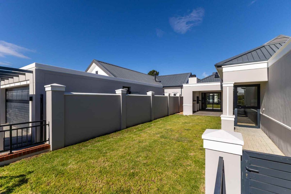 A well-maintained rear exterior area featuring a manicured lawn, paved walkway, secure boundary wall, and modern metal roofing.