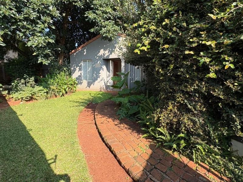 A view of the rear garden featuring a brick pathway leading to a detached structure, surrounded by mature trees and established landscaping.