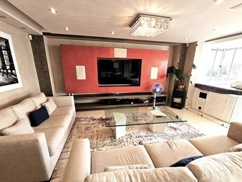 Spacious living room featuring a prominent built-in media wall, recessed ceiling lighting, a decorative crystal chandelier, and neutral tiled flooring.