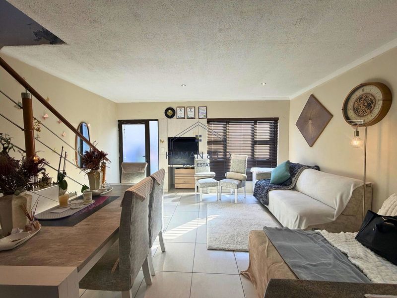 Open-plan living and dining area featuring tiled flooring, a staircase with metal railing, and built-in media cabinetry with natural light from large windows.