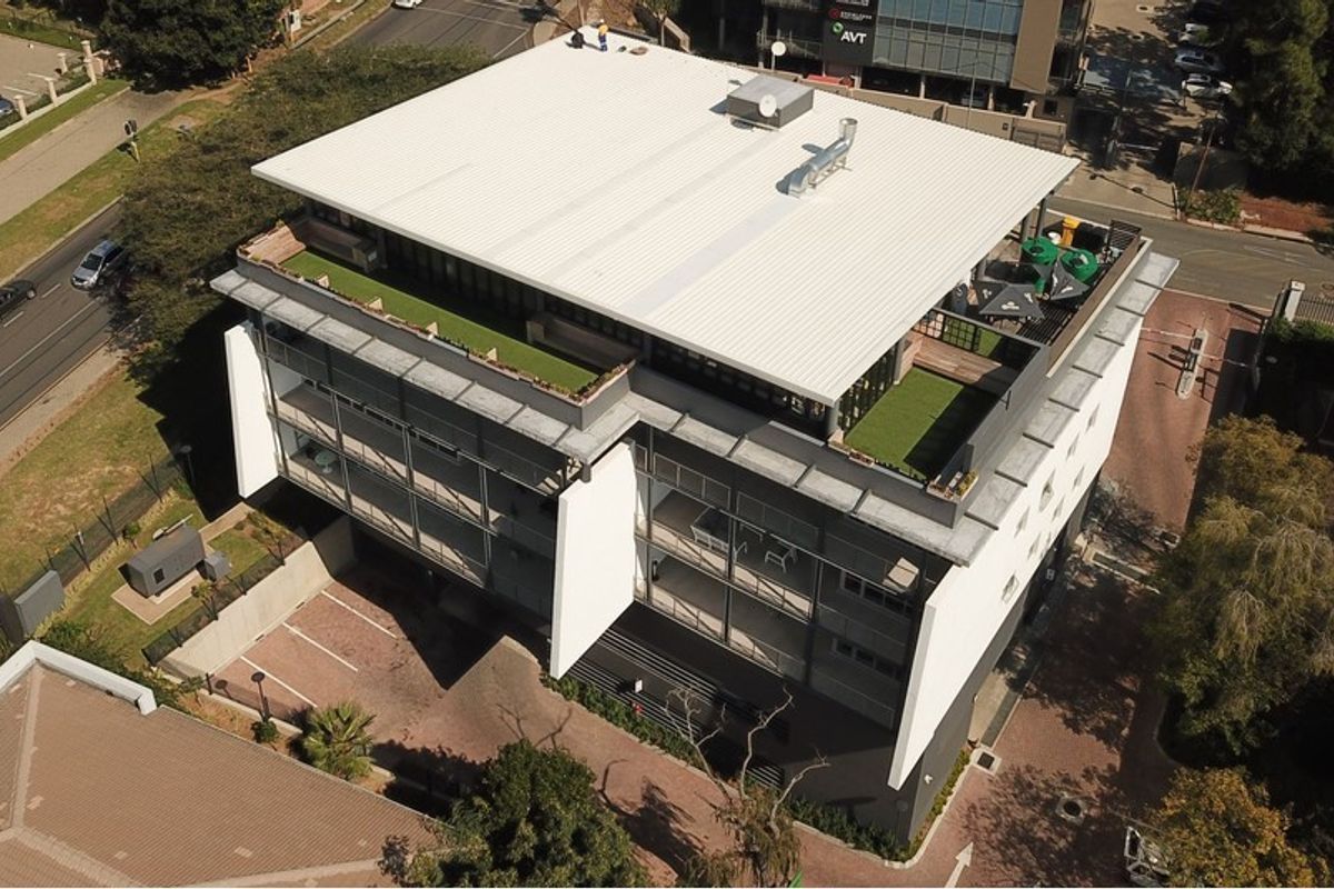 Aerial view of a modern multi-story office building featuring a rooftop terrace with artificial turf and an adjacent paved parking area.