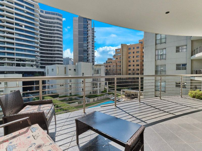 Spacious covered balcony featuring tiled flooring and stainless steel railings, offering an elevated urban view of the surrounding apartment complex and pool area.