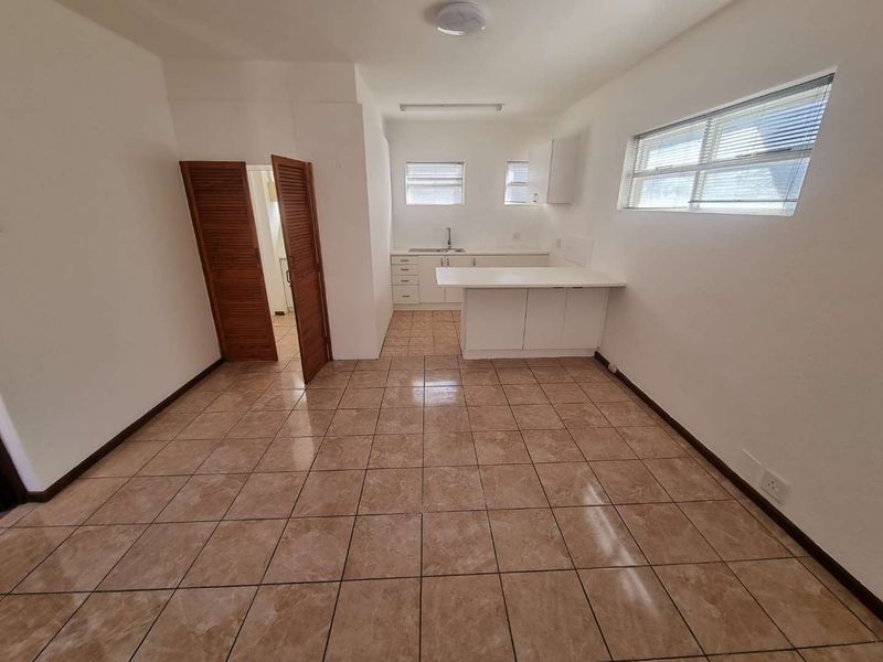 An open-plan living and kitchen area featuring tiled flooring, white cabinetry, a breakfast counter, and louvered doors leading to an adjacent space.