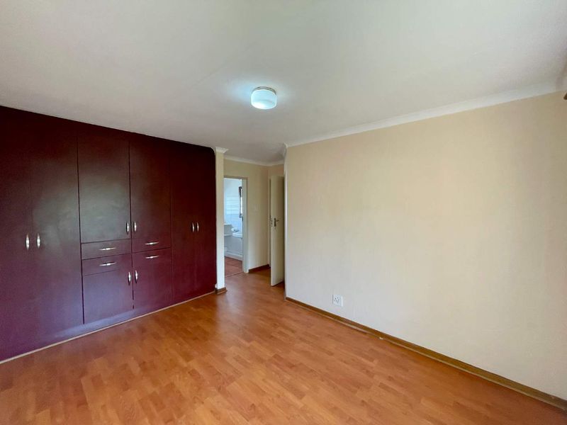 A spacious, empty bedroom featuring dark wood-finish built-in cupboards, laminate flooring, and neutral wall tones, with an adjacent view into a bathroom.