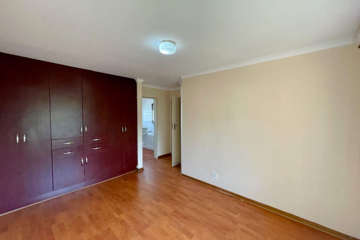 A spacious, empty bedroom featuring dark wood-finish built-in cupboards, laminate flooring, and neutral wall tones, with an adjacent view into a bathroom.