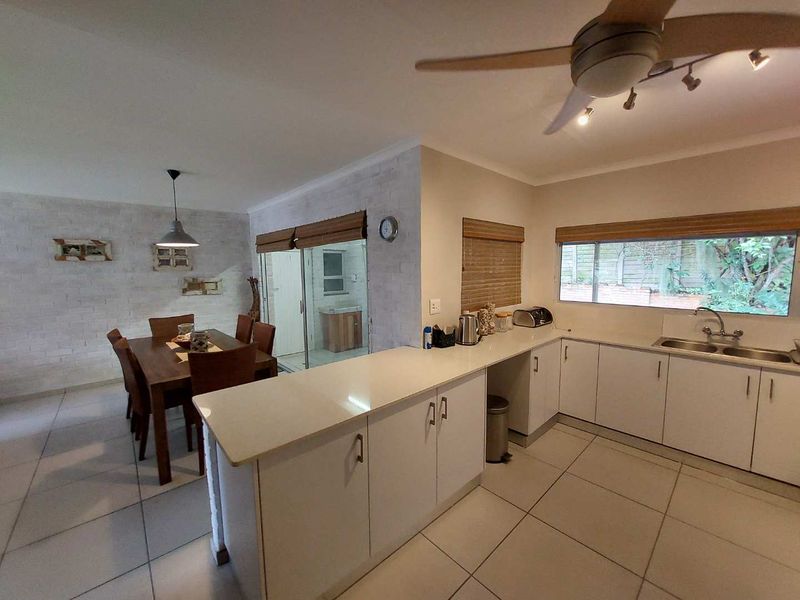 Open-plan kitchen and dining area featuring white cabinetry, tiled flooring, a ceiling fan, and a brick-style feature wall with access to an outdoor space.