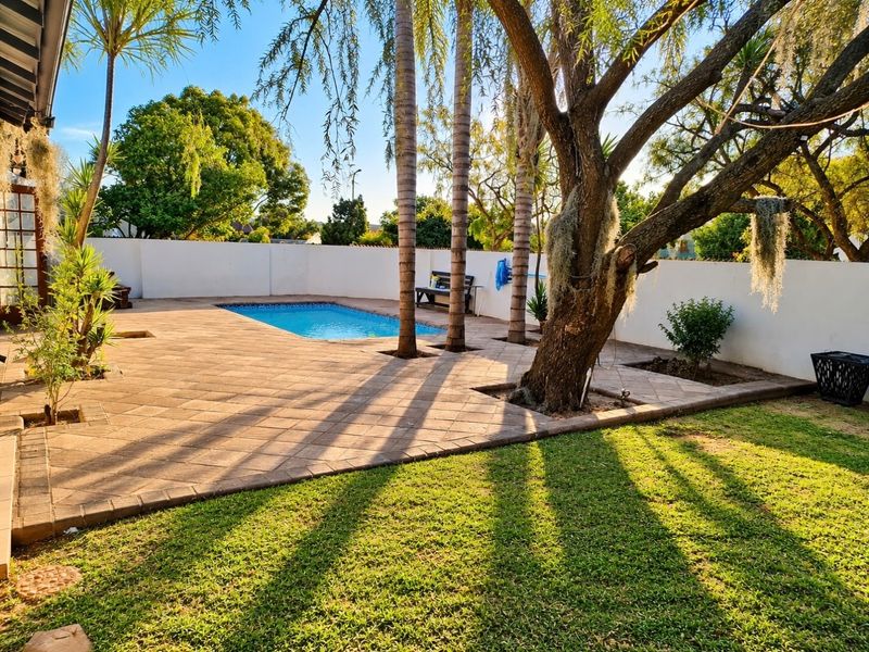 A well-maintained outdoor pool area featuring a paved patio, established trees, and a surrounding boundary wall, perfect for outdoor relaxation.