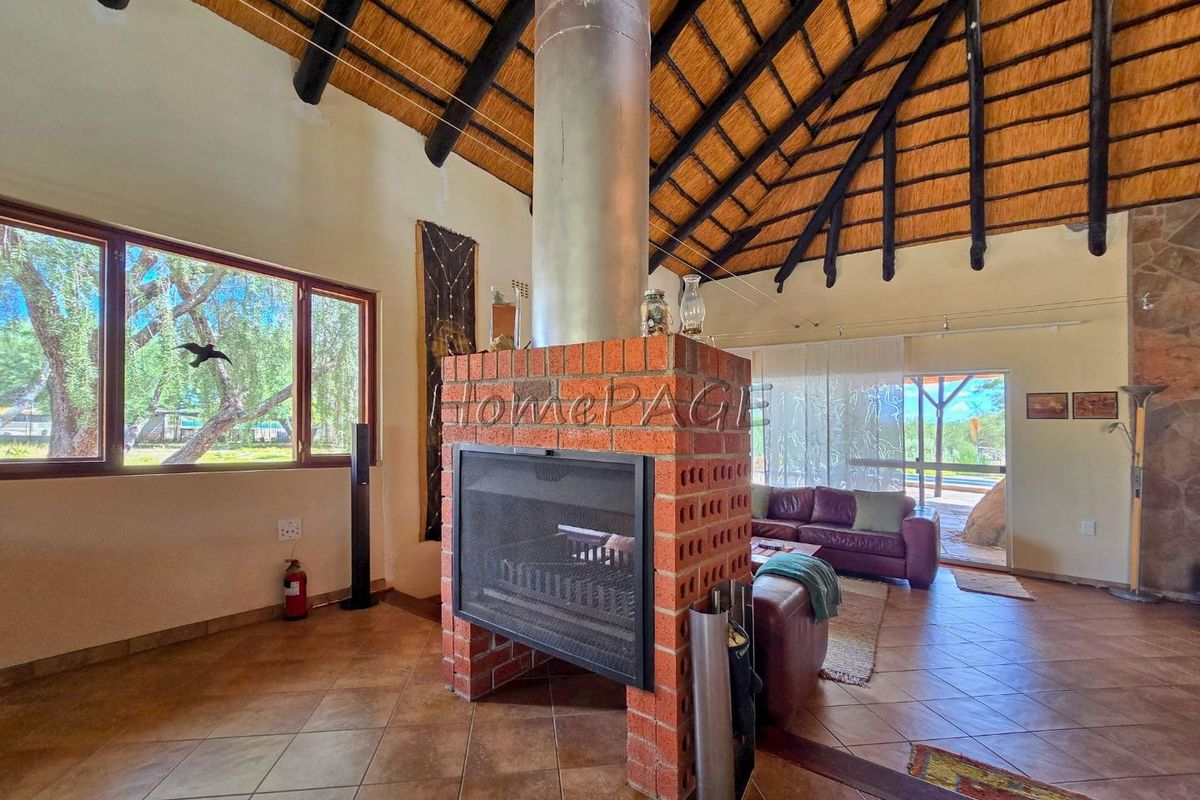 Spacious living area featuring a central exposed brick fireplace, high thatched roof with exposed timber beams, and large windows overlooking the garden.
