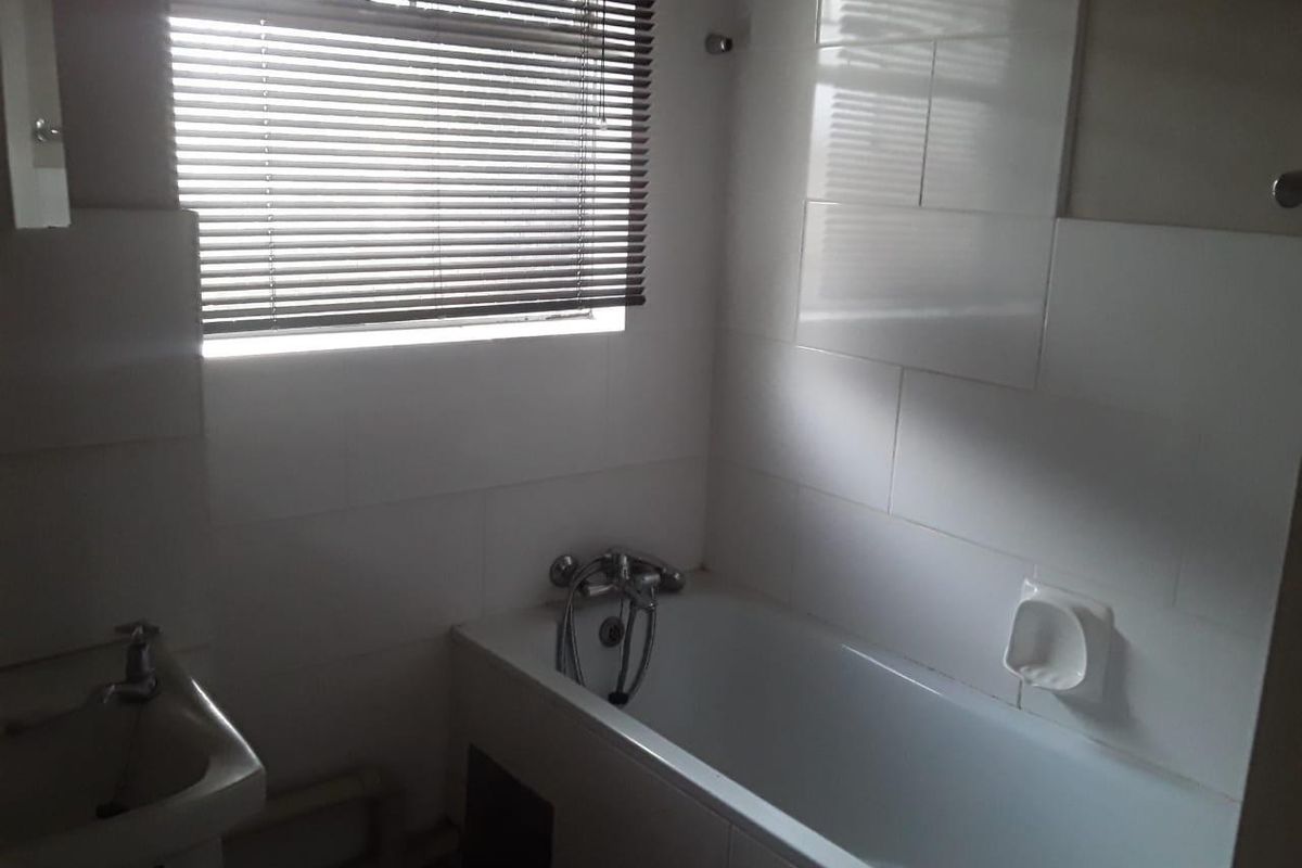 A functional bathroom featuring a white bathtub with shower mixer, wall-mounted basin, and tiled walls with a window blind.