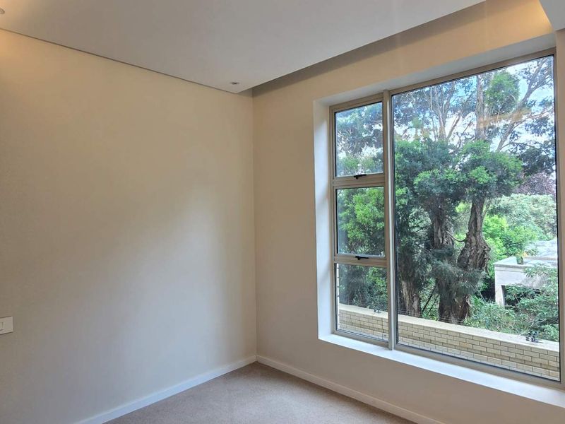 A bright, empty bedroom featuring neutral walls, carpeted flooring, and a large window with a pleasant leafy outlook.