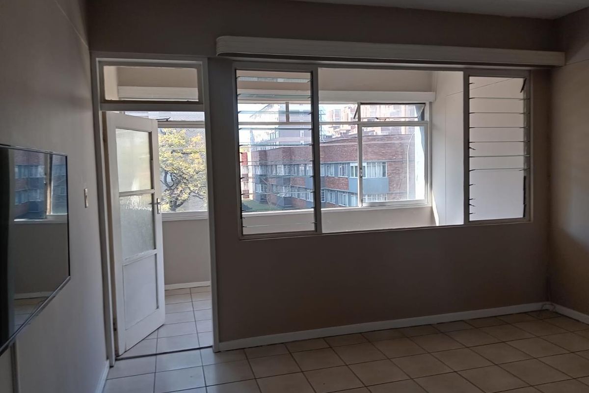 An empty living room featuring tiled flooring, large windows with louvre sections, and direct access to an adjoining balcony area.