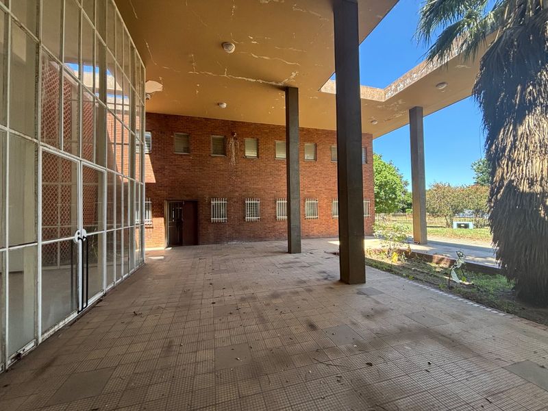 A spacious covered patio area featuring brick walls, concrete support columns, and large glass windows, showing signs of wear and requiring maintenance.