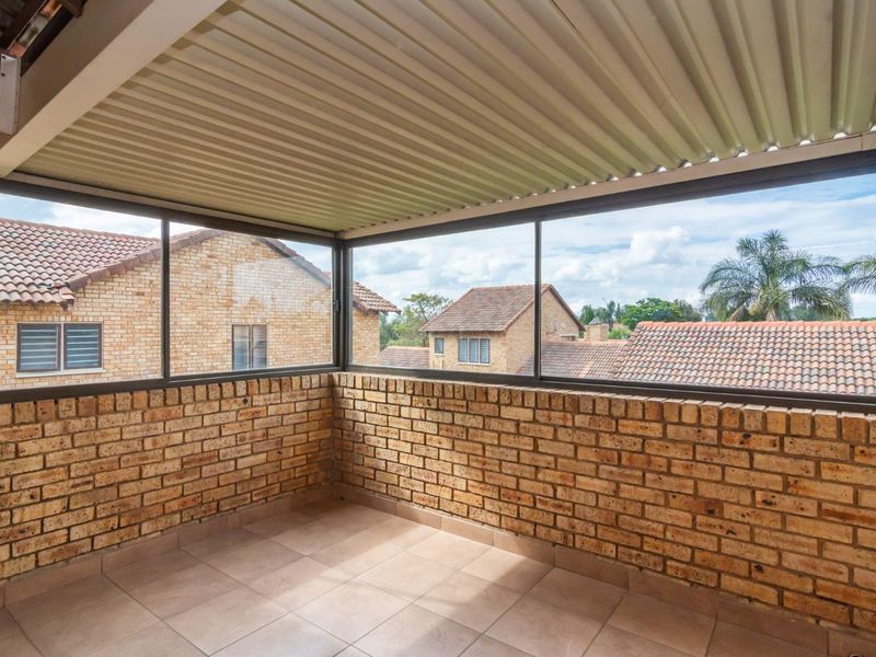 A covered balcony featuring exposed brick walls, tiled flooring, and large aluminium-framed windows offering an elevated view of the surrounding residential complex.