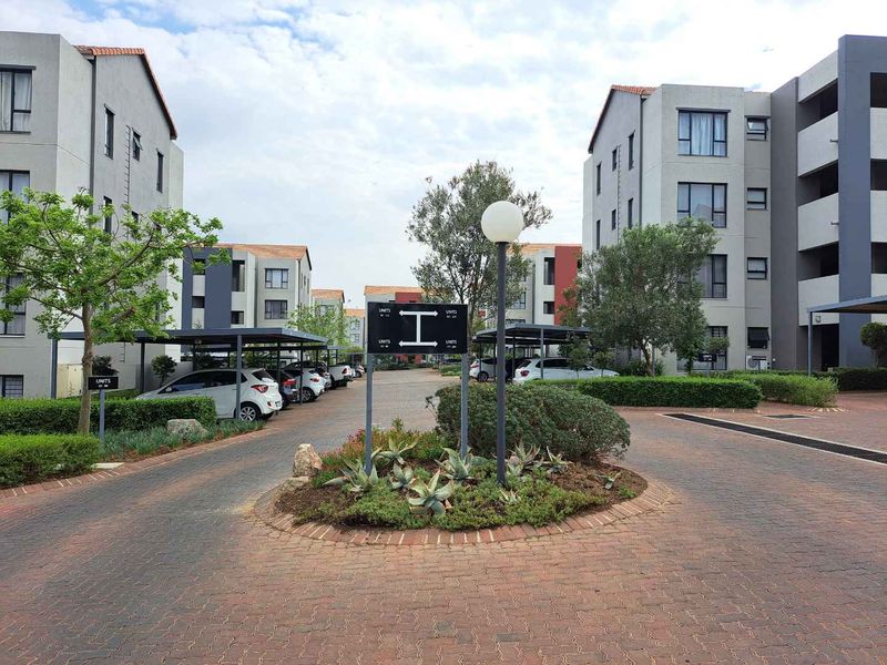 A well-maintained apartment complex courtyard featuring paved driveways, covered parking bays, and established landscaped garden beds between multi-story residential buildings.