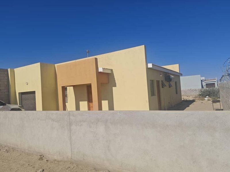 Front view of a single-story house featuring a yellow facade, integrated garage, and a concrete boundary wall.