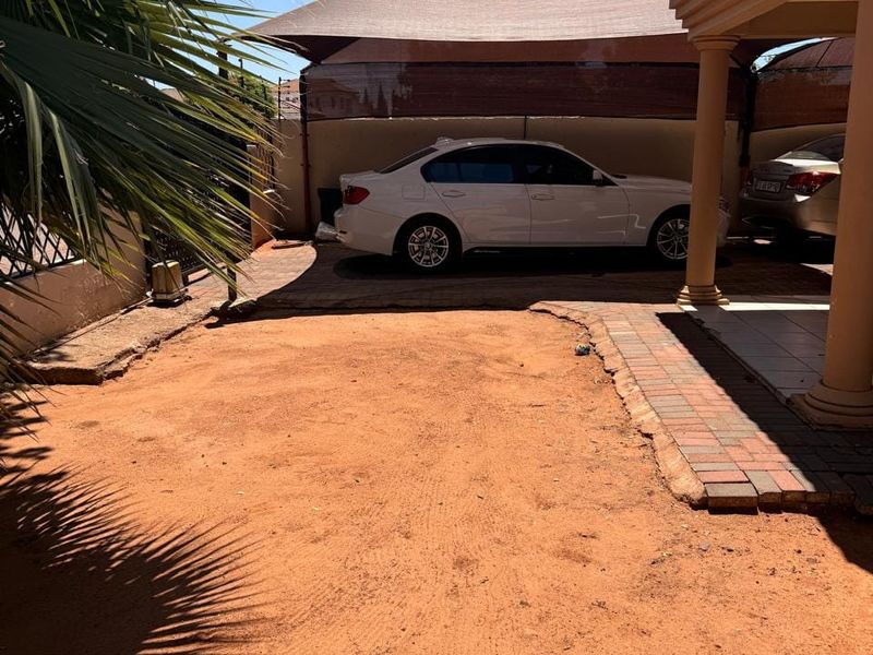 Front exterior area featuring a covered porch with columns and a shade cloth carport structure over a paved and sandy driveway section.