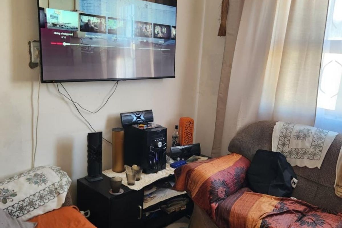 A living room area featuring a wall-mounted television and window with curtain rail, showing signs of wear on the wall surfaces.