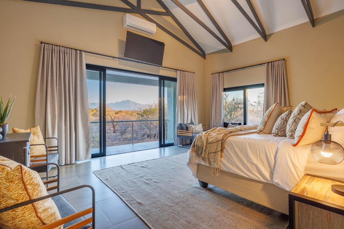 Spacious bedroom featuring vaulted ceilings with exposed rafters, tiled flooring, and large sliding glass doors opening onto a private balcony with scenic views.
