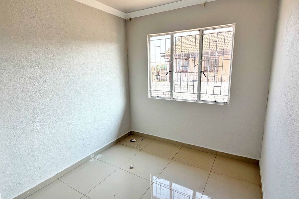A bright, empty bedroom featuring neutral tiled flooring, white walls, and a window fitted with security bars.