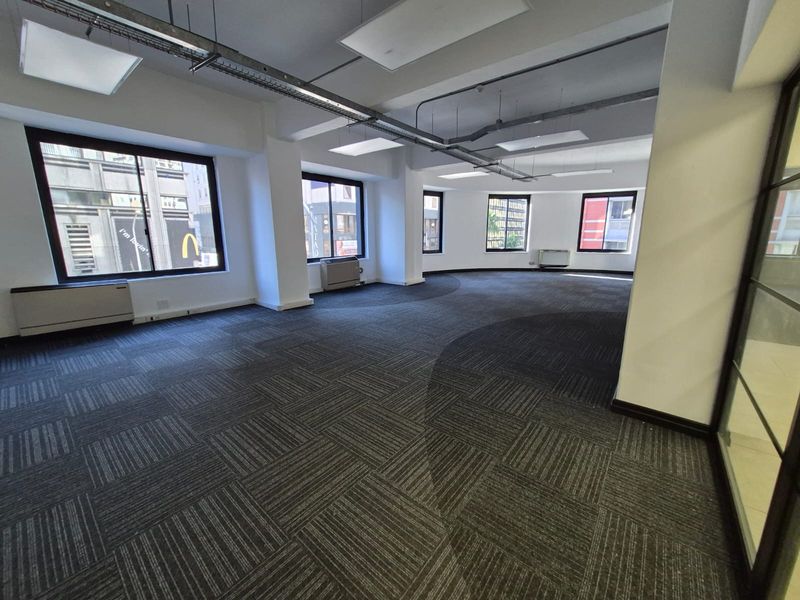 Spacious open-plan office area featuring carpet tiles, exposed ceiling services, and large windows providing ample natural light.